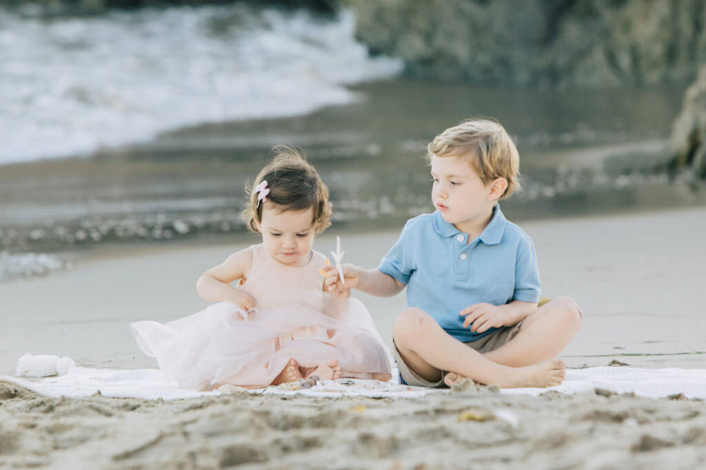 two kids seating on the beach wearing pastel tones 