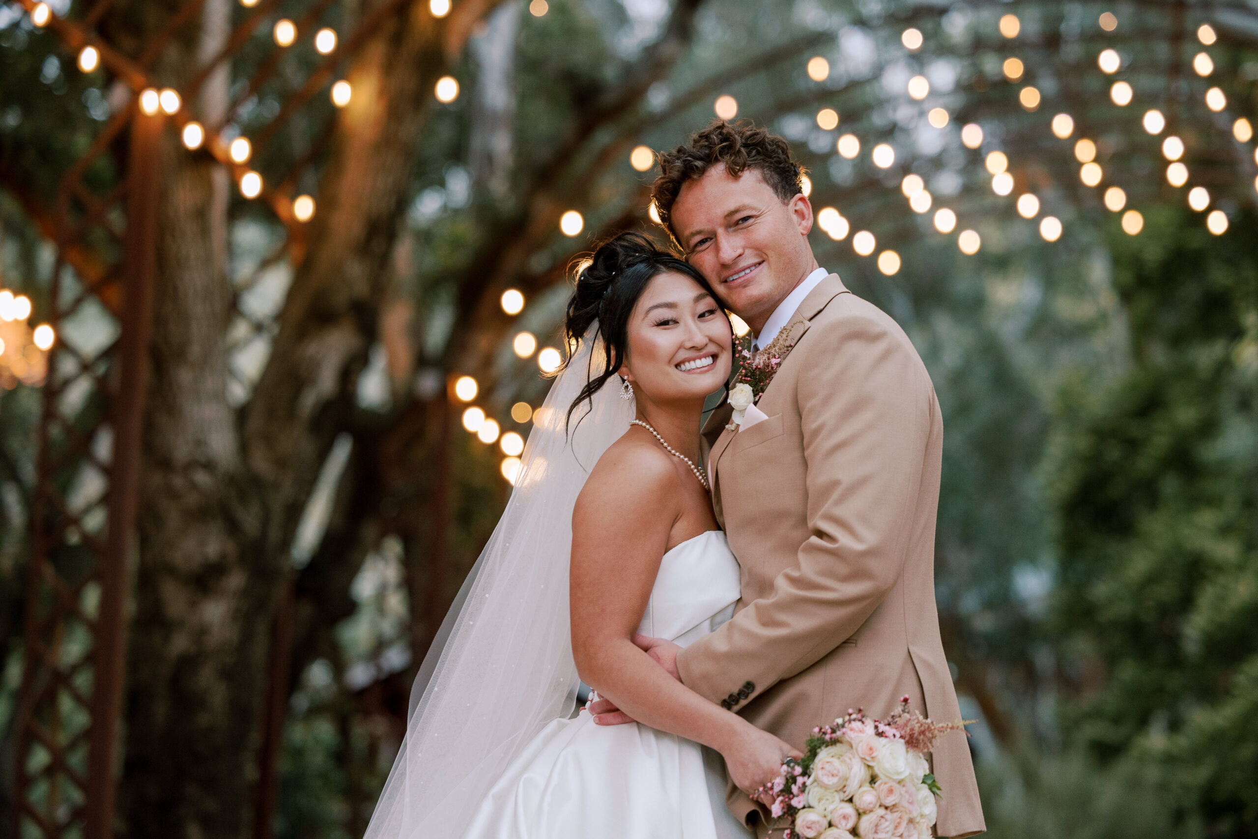Groom wearing a tan suit hugging his bride holding a blush pink bridal bouquet at Calamigos Ranch