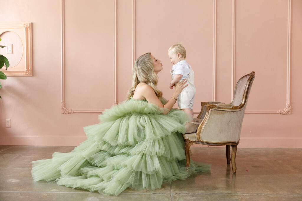 Elegant studio portrait of a mother in a green tulle dress holding her baby against a soft pink wall with a vintage chair.