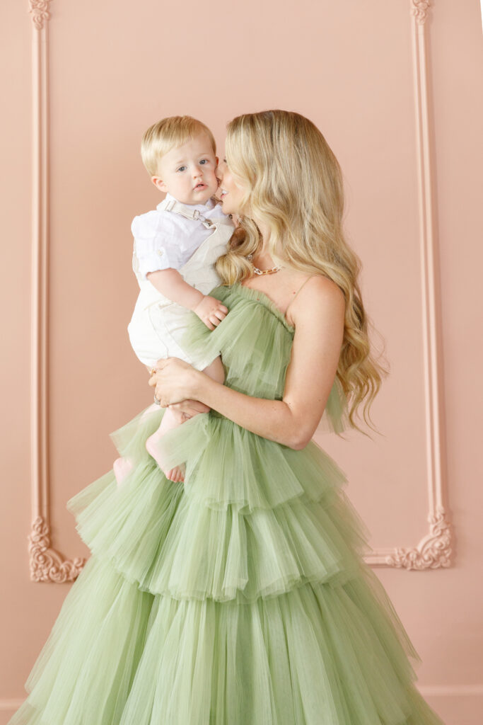 Mother holding her baby during an elegant studio family portrait session with a soft pink backdrop and green tulle gown