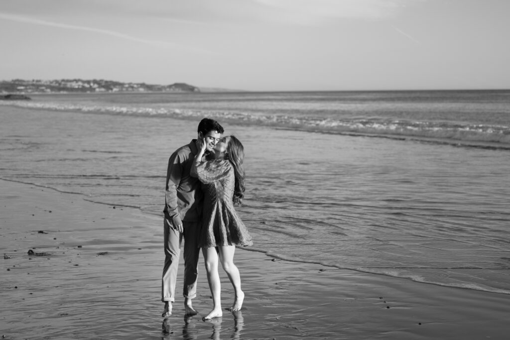 Black-and-white portrait of a couple sharing a playful kiss along the shoreline