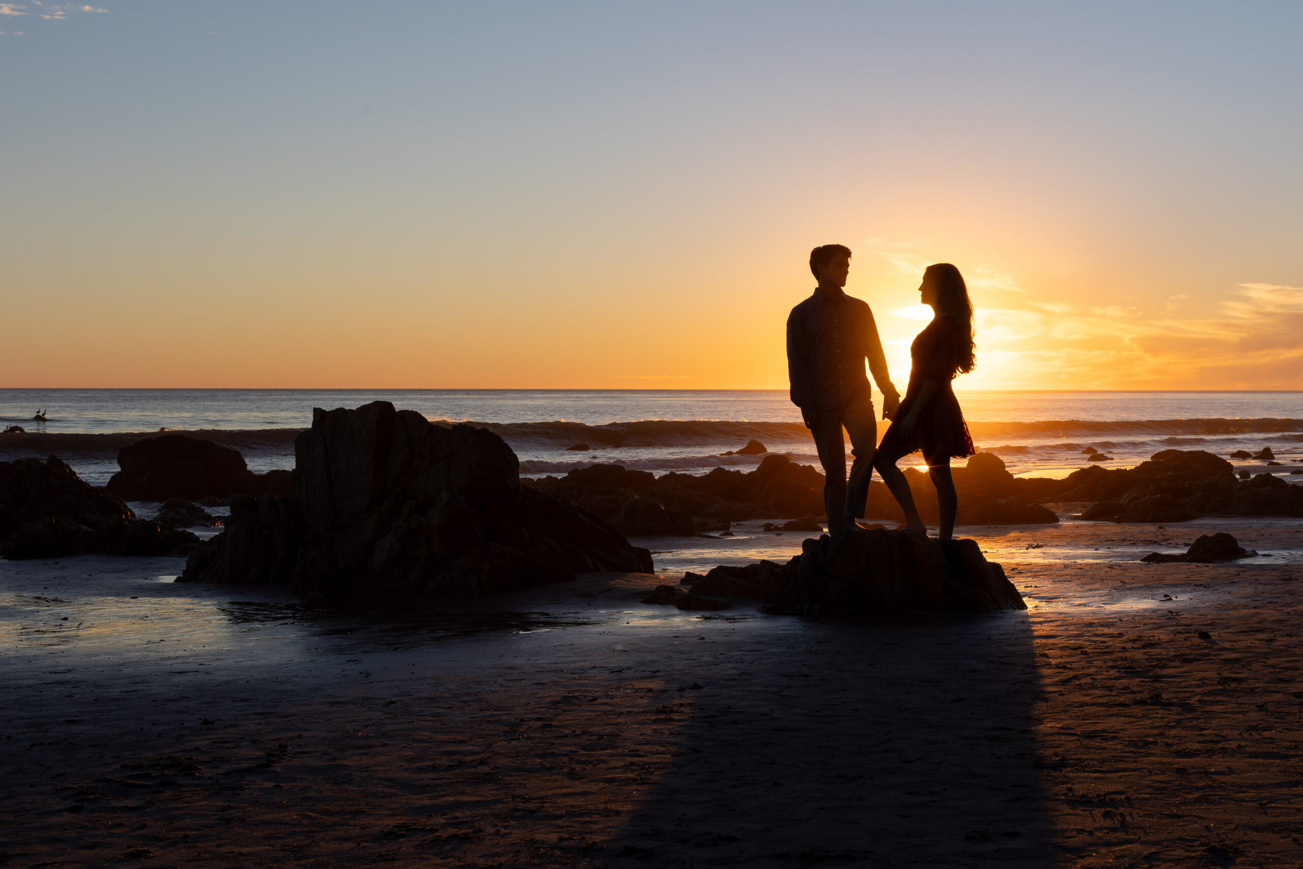 Silhouette of an engaged couple holding hands on beach rocks at sunset.