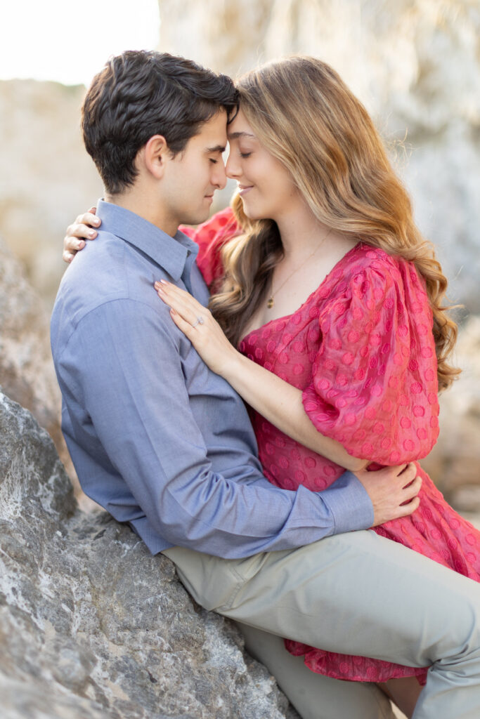 Engaged couple touching foreheads while seated together on coastal rocks