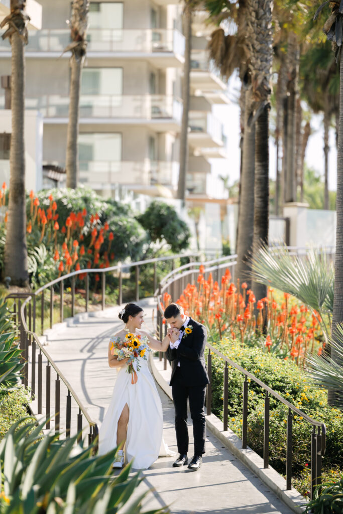 Groom kissing the bride’s hand while walking through tropical landscaping near their Huntington Beach wedding venue.