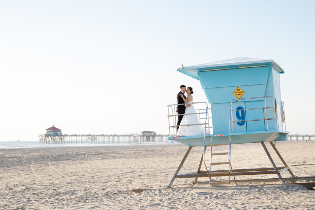 Bride and groom photographed beside an iconic blue Huntington Beach lifeguard tower with the pier in the distance.