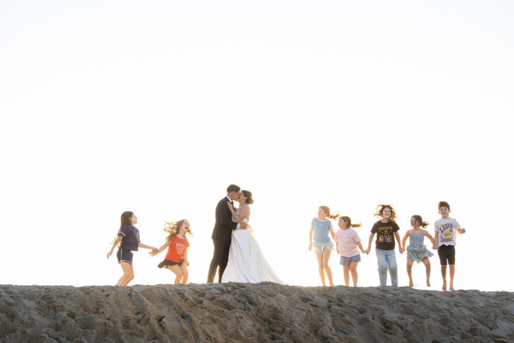 Bride and groom kissing as children play around them during a joyful candid moment on Huntington Beach