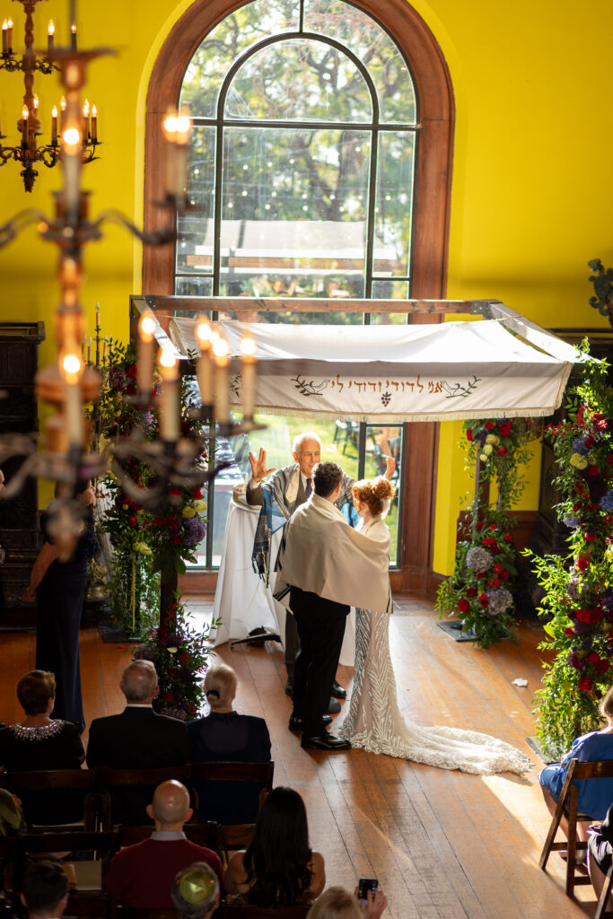 Bride and groom wrapped together in a tallit during their Jewish wedding ceremony