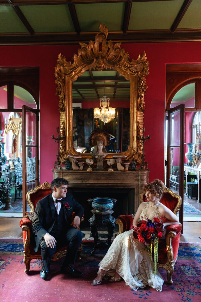 Bride and groom seated in vintage red chairs inside one of Paramour Estate’s dramatic rooms