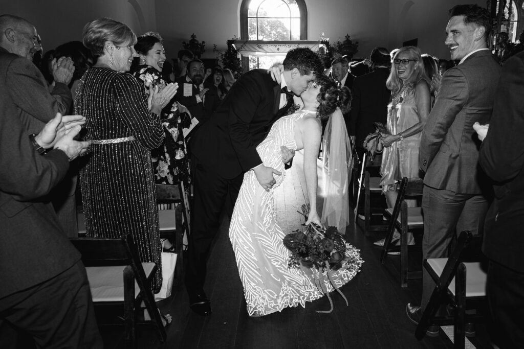 oyful black-and-white photograph of the newlyweds kissing in the aisle after their ceremony