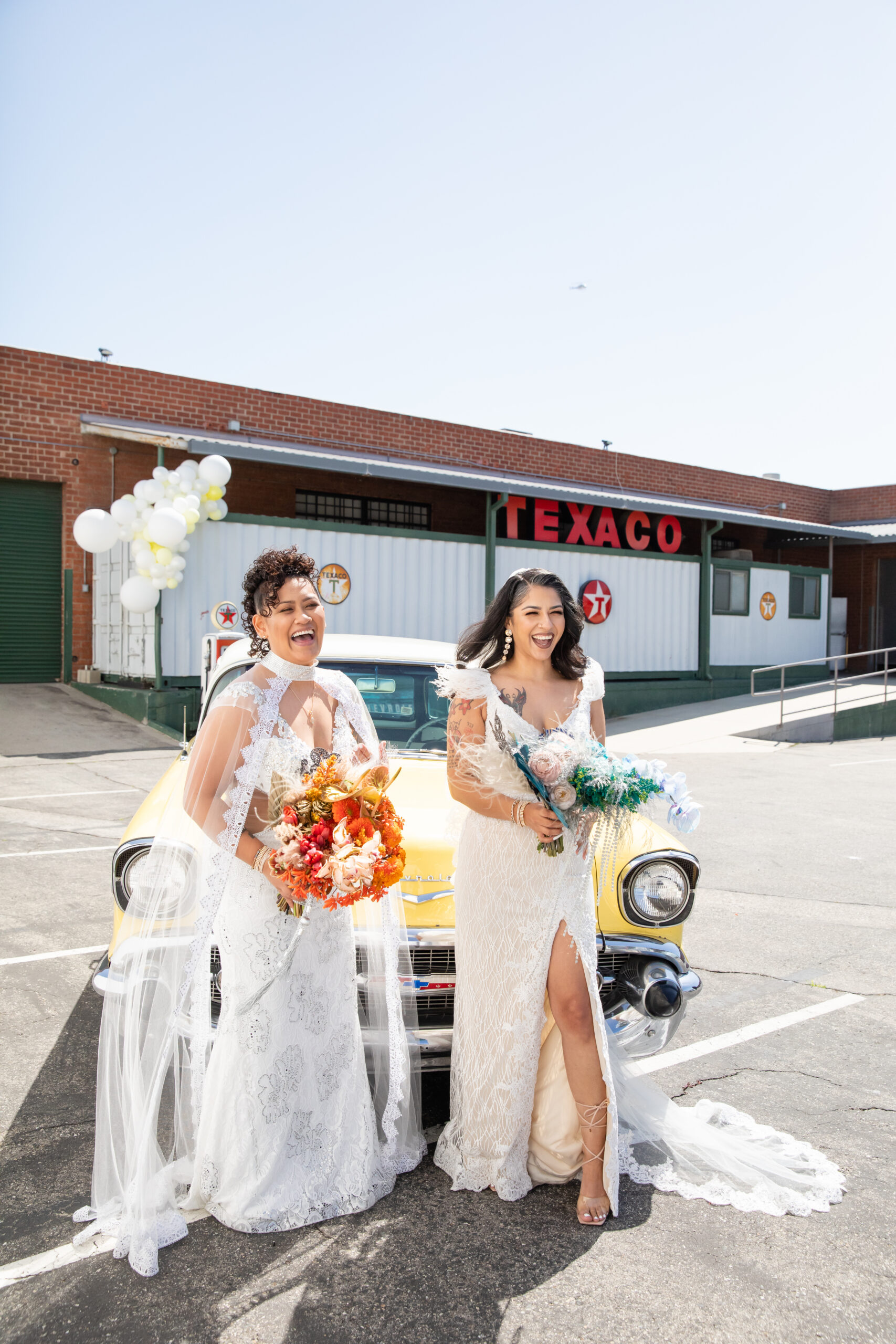 Two brides laughing together in front of a yellow vintage car at their Los Angeles wedding