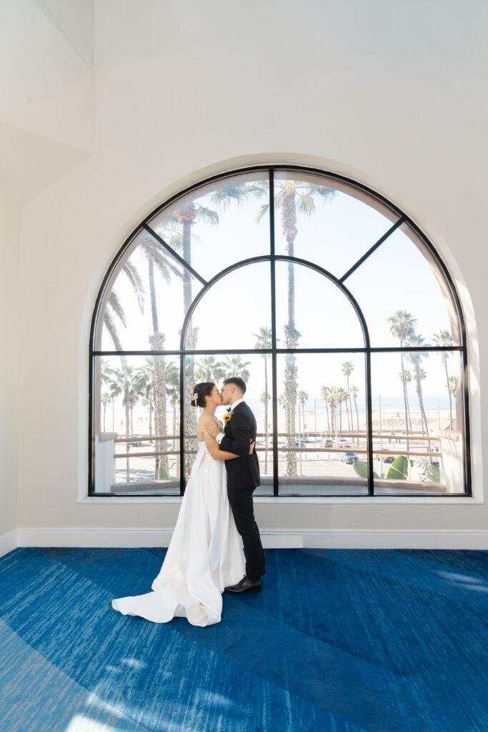 Bride and groom kissing beneath the large arched window overlooking Huntington Beach and the Pacific Ocean.