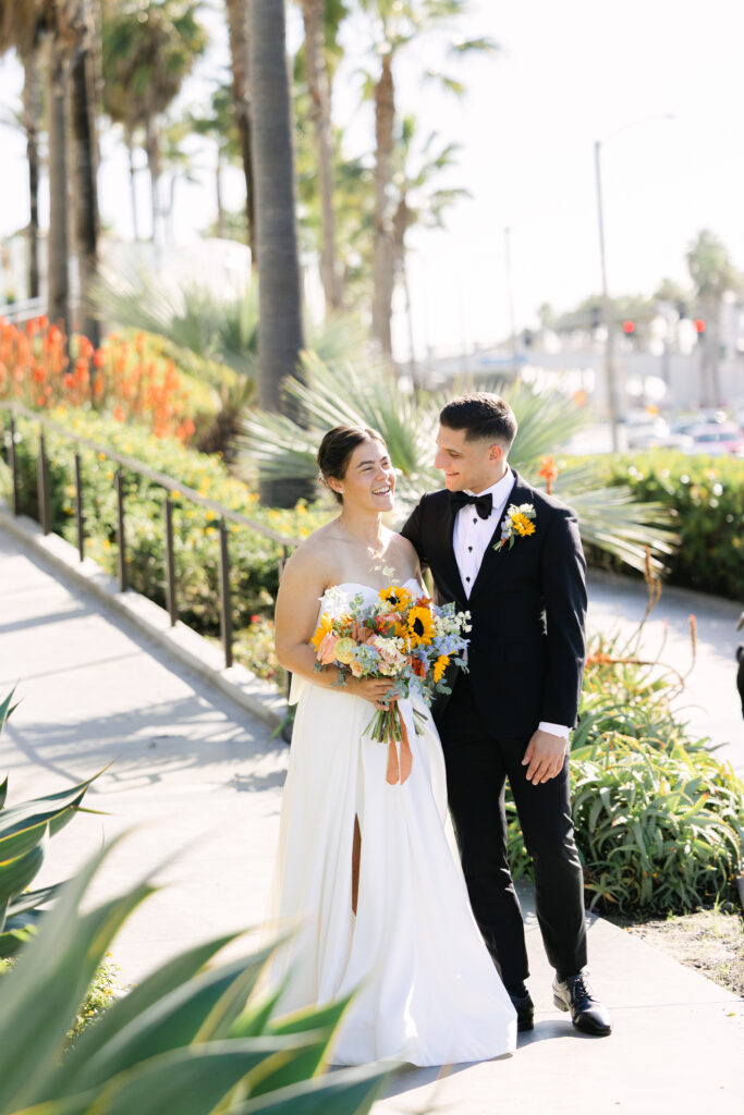 Laughing bride and groom enjoying a relaxed portrait together surrounded by palm trees and colorful flowers.